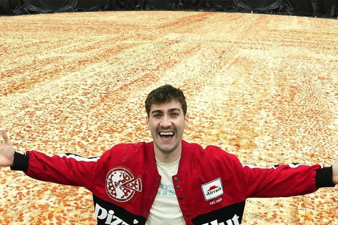 Uśmiechnięty mężczyzna w czerwonej kurtce prezentuje największą pizzę świata, która rozciąga się aż po horyzont.