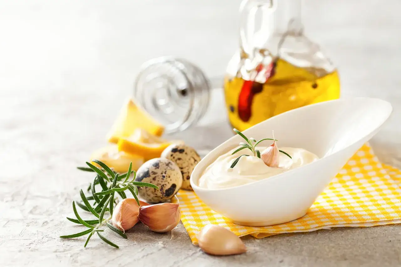 Kremowe aioli w białej miseczce, ozdobione gałązką rozmarynu i ząbkiem czosnku. Obok składniki: jajka przepiórcze, czosnek, cytryna, oliwa.