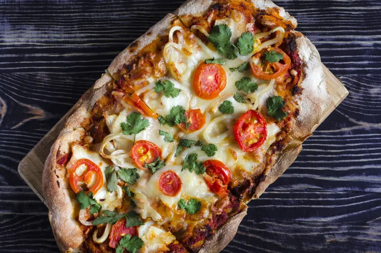 Pinsa co to? Aromatyczna, prostokątna pizza z pomidorkami, cebulą i serem, udekorowana świeżą kolendrą, na drewnianym tle.