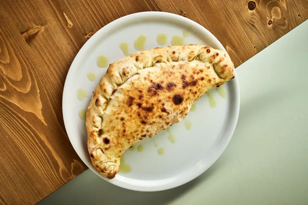 Złocisty calzone, czyli pieczona włoska kieszonka z ciasta, lśni na białym talerzu, skropiony oliwą.