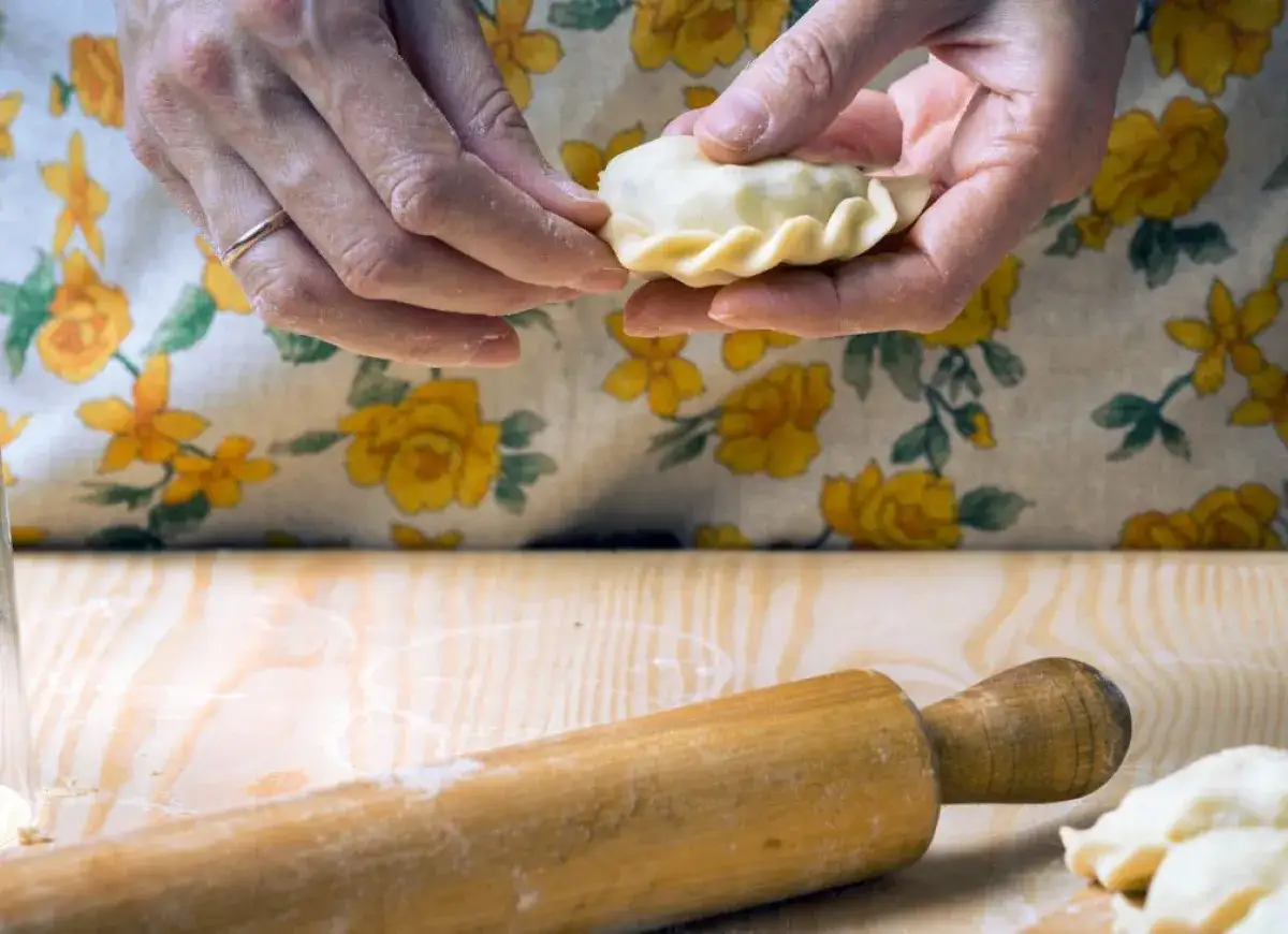 Dłonie formują pieroga, który będzie częścią przepisu na miękkie ciasto na pierogi. Obok wałek i mąka.