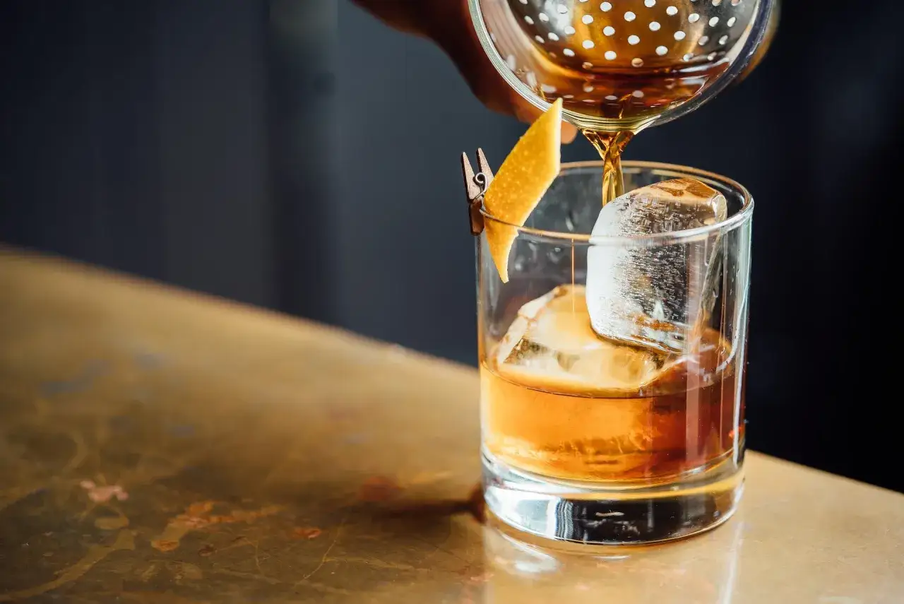 Jak zrobić krystaliczny lód w domu? Barman przelewa bursztynowy trunek do szklanki z dużym, przejrzystym lodem i skórką pomarańczy.