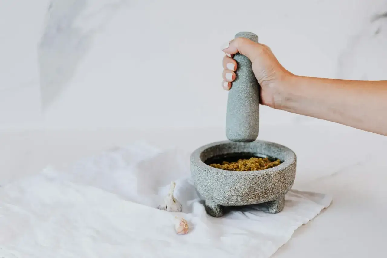 Ręka uciera w moździerzu świeże zioła, tworząc aromatyczne pesto z bazylii. Obok leżą ząbki czosnku.