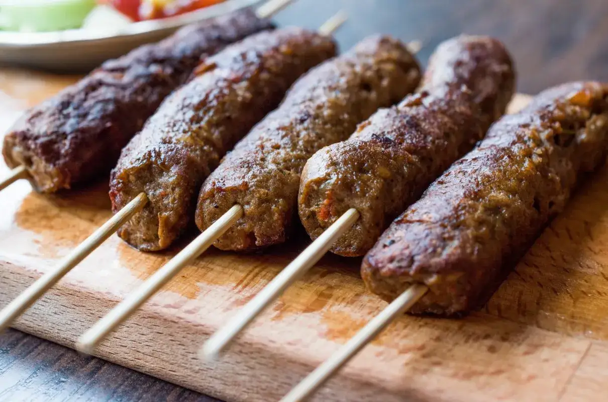 Pięć soczystych kofta kebabów na drewnianych patyczkach, ułożonych na desce. Idealne na grilla.