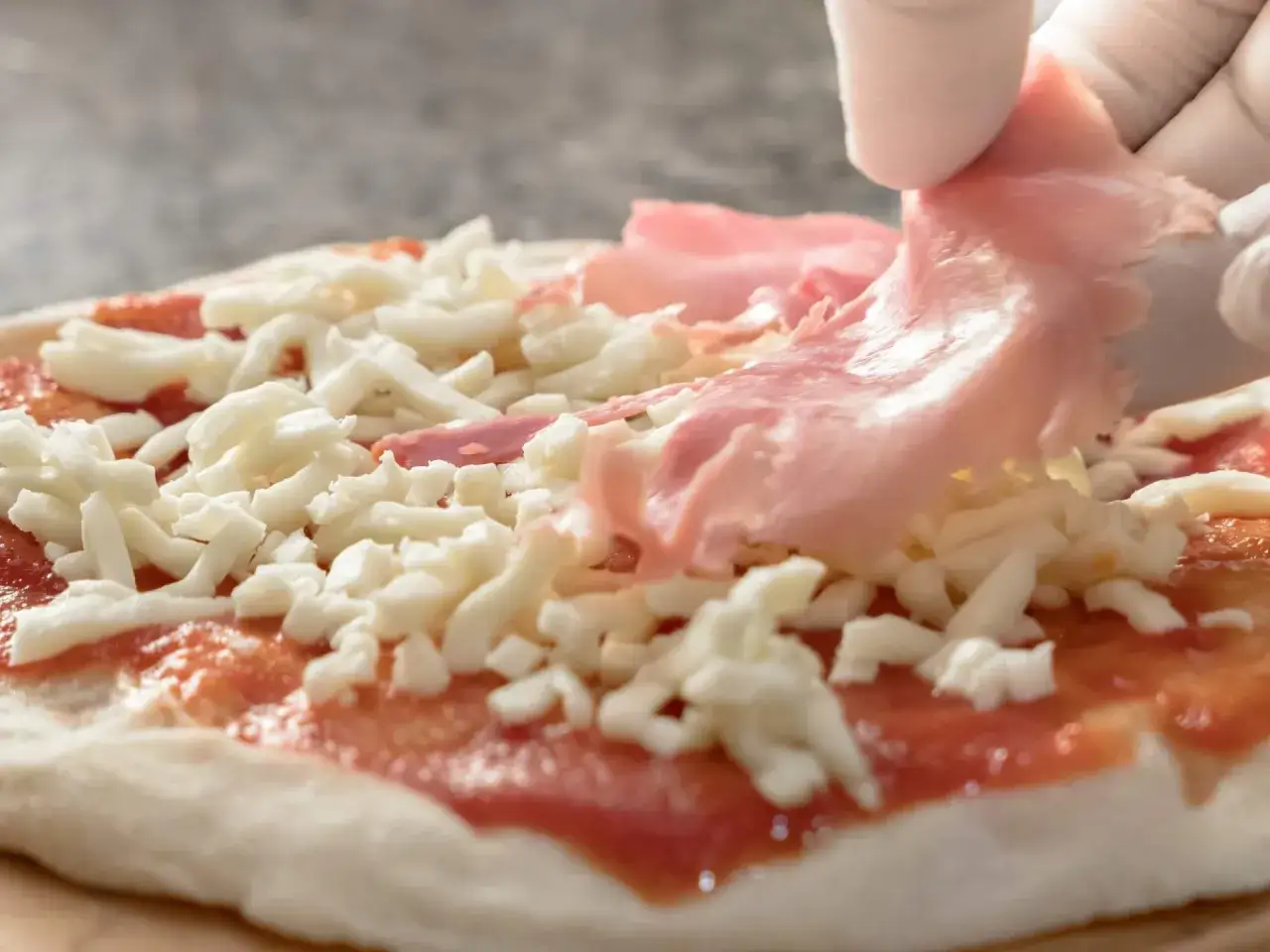 Rękawiczki zakładają cienkie plastry różowej szynki na pizzę z serem i sosem pomidorowym.